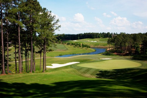 A scenic golf course with lush green fairways, a winding river, and sand traps under a blue sky with fluffy clouds.