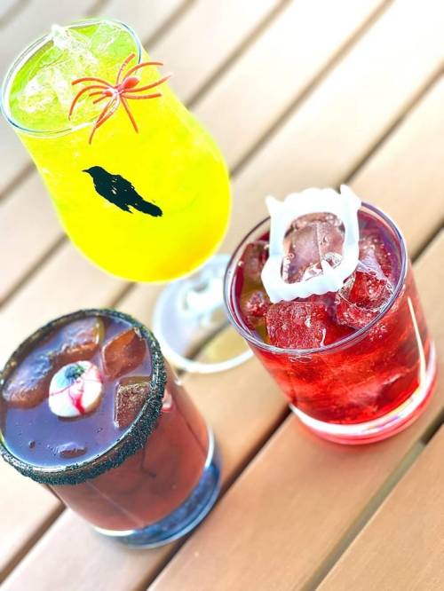 Three colorful cocktails on a wooden table: a yellow drink with a spider garnish, a dark drink with a cherry, and a red drink with ice.