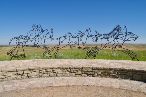 A metal sculpture depicting a historical scene of figures on horseback against a clear blue sky and grassy landscape.