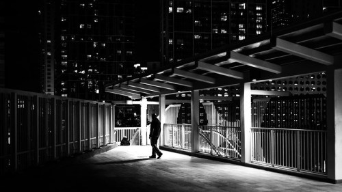 A solitary figure walks on a rooftop at night, illuminated by city lights and shadows in a black and white scene.