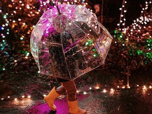 A person walks under a clear umbrella, surrounded by colorful lights and rain, wearing yellow boots.