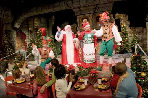 A festive scene with Santa, Mrs. Claus, and performers in a decorated room, while families enjoy a meal at a table.