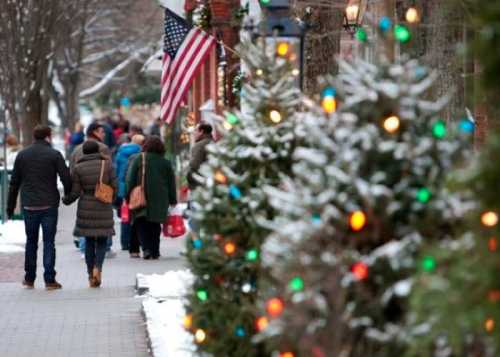 A couple walks hand in hand along a snowy street, decorated with colorful lights and holiday shoppers in the background.