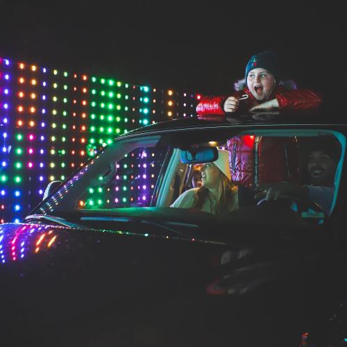 A child in a red jacket excitedly leans out of a car window, with colorful lights glowing in the background.