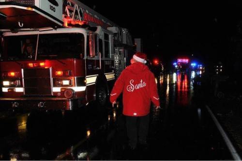 A person in a red "Santa" jacket walks towards emergency vehicles on a wet road at night.