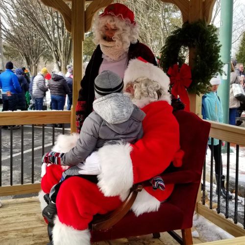A child sits on Santa's lap while Mrs. Claus stands nearby, all in a festive outdoor setting.