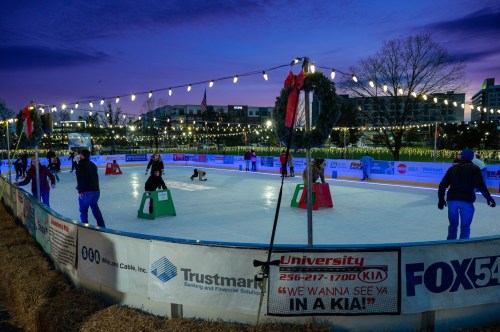 Outdoor ice skating rink at dusk, decorated with lights and festive bows, with skaters enjoying the activity.