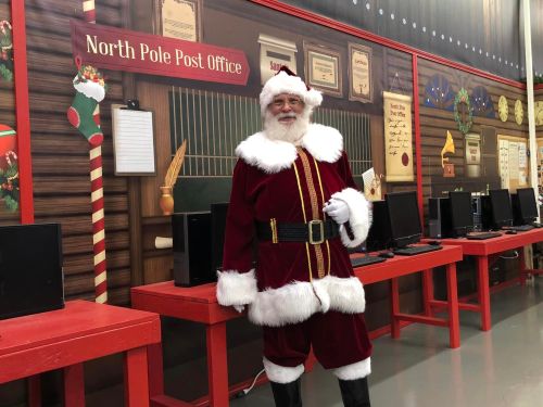 Santa Claus stands in a festive North Pole Post Office, surrounded by red tables and computers, smiling cheerfully.