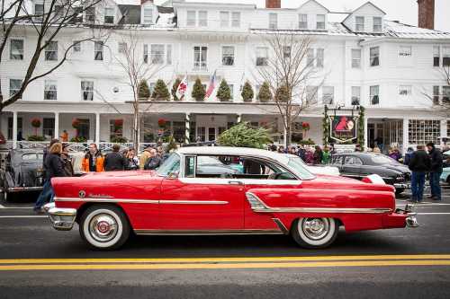 A classic red car parked on a street in front of a historic building, with a crowd of people nearby.