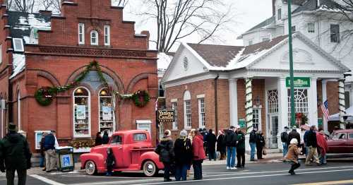 A festive street scene with a vintage pink truck, historic buildings, and a crowd of people enjoying the holiday atmosphere.