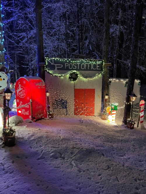A whimsical, snowy scene featuring a decorated post office with festive lights and a red mailbox.