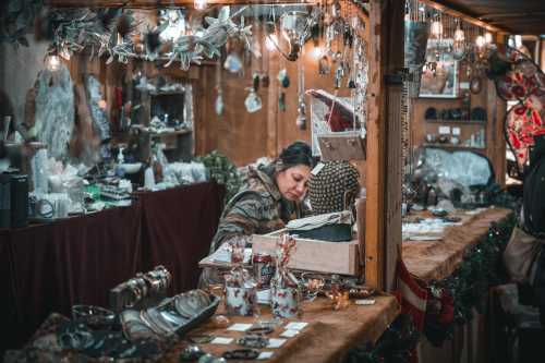 A woman examines items at a cozy market stall filled with various crafts and decorations. Warm lighting enhances the atmosphere.