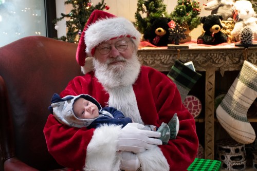 Santa Claus holds a smiling baby in a cozy setting decorated for Christmas, with festive greenery and stockings in the background.