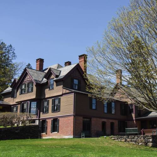 A large, historic house with multiple gables, surrounded by trees and a grassy lawn on a sunny day.