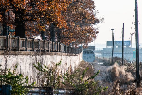 A weathered concrete wall lined with trees in autumn, leading to a distant industrial area under a hazy sky.