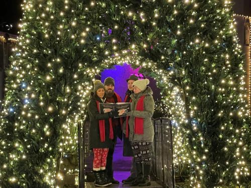 A group of three people stands under a lit archway, wearing winter clothing and holding a sign, surrounded by festive decorations.