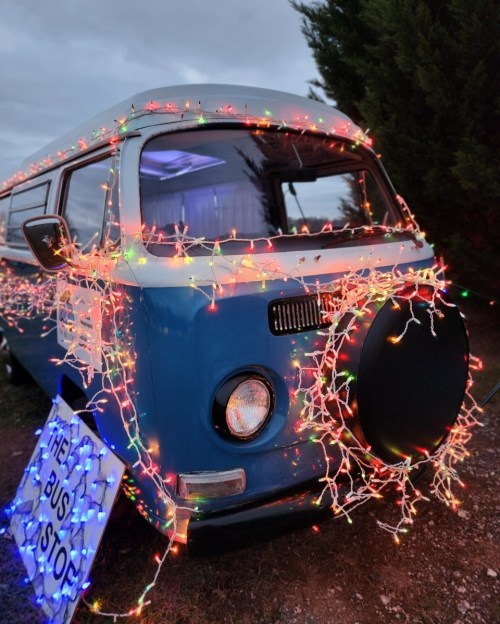 A vintage blue van decorated with colorful Christmas lights, parked beside a sign that reads "The Bus Stop."