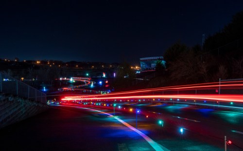 A night scene featuring colorful light trails along a winding road, with distant city lights and trees in the background.