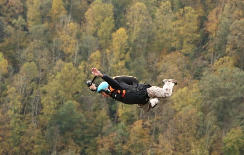 A skydiver in a black suit and blue helmet leaps through the air above a forest of autumn-colored trees.