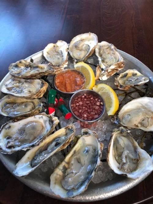 A platter of fresh oysters on ice, accompanied by lemon wedges, cocktail sauce, and diced onions.