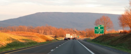 A highway scene with cars, a sign for Shenandoah Caverns, and mountains in the background during sunset.