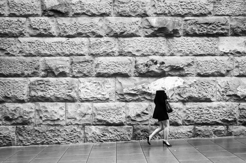 A person in a black dress and heels walks with an umbrella against a textured stone wall in black and white.