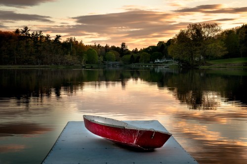 A red boat rests on a dock at sunset, reflecting in calm water surrounded by trees and a serene landscape.