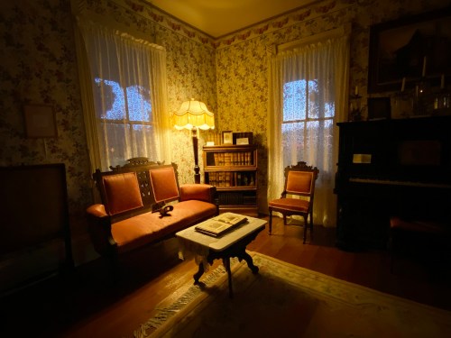 Cozy vintage room with floral wallpaper, a sofa, chairs, a lamp, and a piano, softly lit by warm light.