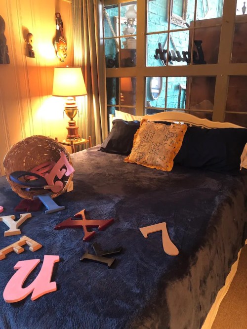 A cozy bedroom with a blue bedspread, decorative pillows, and colorful letters scattered on the bed. A lamp illuminates the space.