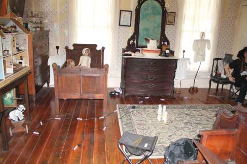 A vintage room with wooden furniture, a mirror, dolls on a bed, and scattered items on the floor.