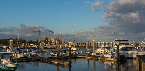 A marina filled with boats, with a backdrop of clouds and a distant industrial building under a blue sky.