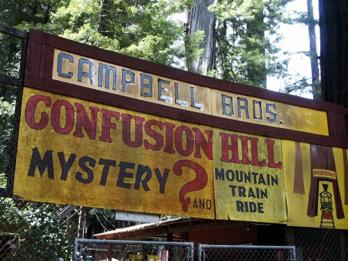 Sign for "Campbell Bros. Confusion Hill" featuring "Mystery?" and "Mountain Train Ride" amidst redwood trees.