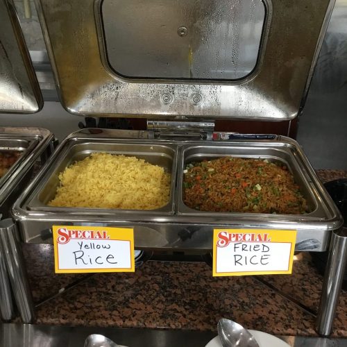 Two metal trays of rice: one with yellow rice and the other with fried rice, both labeled on a buffet table.