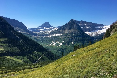 A panoramic view of a mountainous landscape with green valleys, snow-capped peaks, and a clear blue sky.