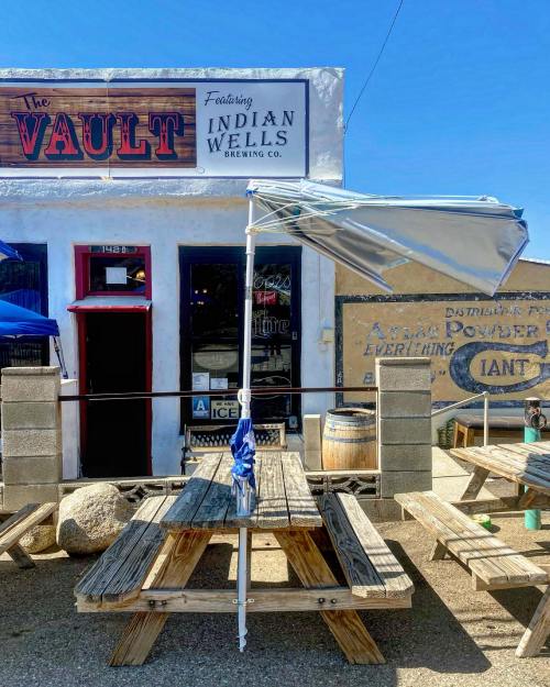 A sunny outdoor seating area with wooden picnic tables and an umbrella in front of a building featuring a sign for "The Vault."