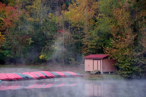 A serene lakeside scene with a small cabin and canoes, surrounded by colorful autumn foliage and morning mist.