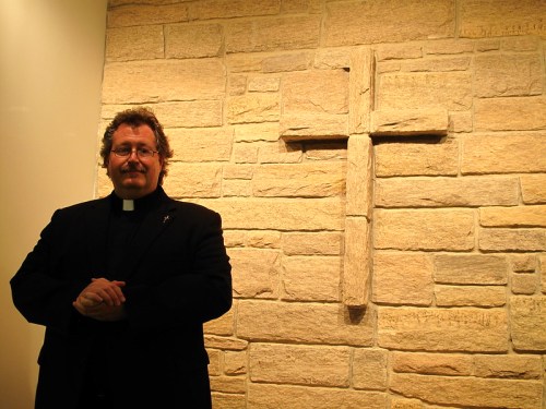 A man in a black suit stands beside a stone wall featuring a cross. He has a friendly expression and is clasping his hands.