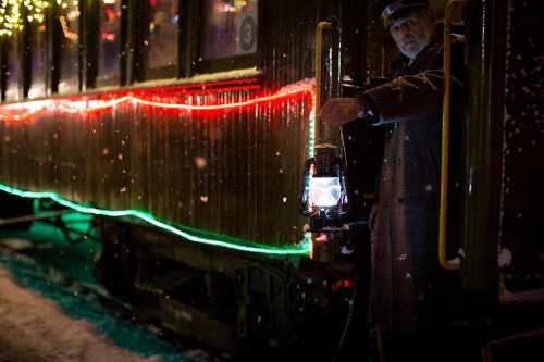 A train conductor holds a lantern by a decorated train, illuminated with colorful lights in a snowy setting.