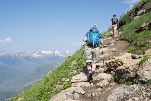 Two hikers ascend a rocky trail with mountains in the background under a clear blue sky.
