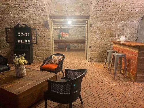 Cozy interior of a stone-walled room with a wooden bar, seating area, and decorative elements. Warm lighting enhances ambiance.