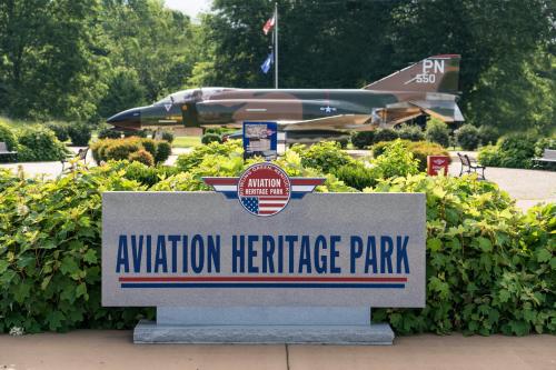 Sign for Aviation Heritage Park with a military jet displayed in the background, surrounded by greenery and flags.