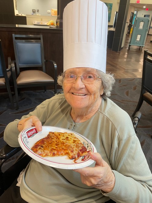 An elderly woman in a chef's hat smiles while holding a plate of pizza.