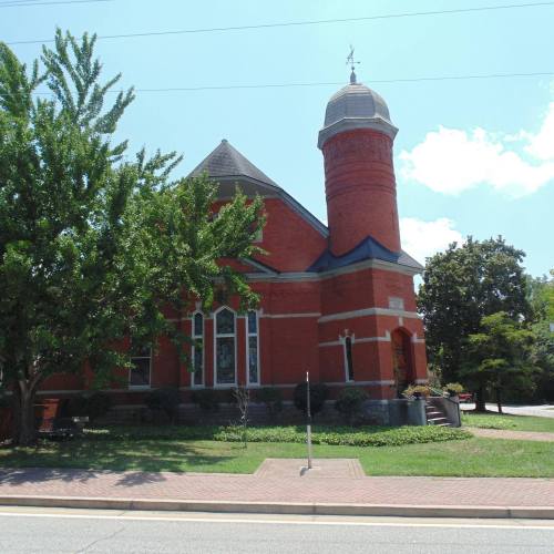 A red brick church with a tall tower, surrounded by trees and grass, on a sunny day.