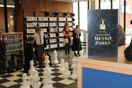 A sign for Louisville Metro Parks in a library, with children playing chess on a large board in the background.