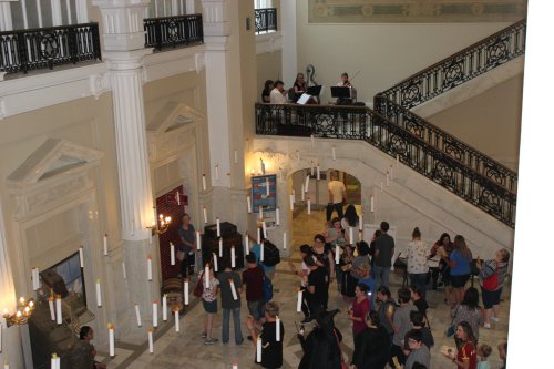 A busy indoor space with people gathered, musicians playing on a balcony, and candles hanging from the ceiling.