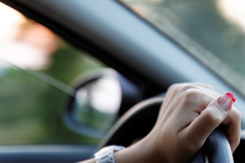 A close-up of a hand with red nails gripping a steering wheel inside a car, with a blurred background.
