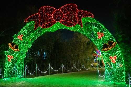 A festive archway adorned with red and green lights, featuring a large bow, holly, and bells, set against a night sky.