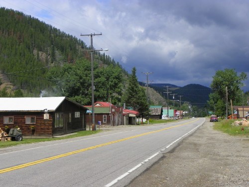 A quiet rural road lined with small buildings and trees, set against a backdrop of mountains and cloudy skies.