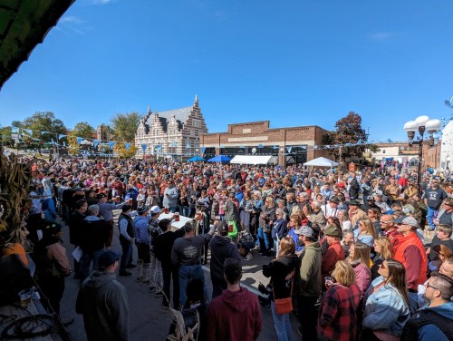 A large crowd gathers at an outdoor event under a clear blue sky, with festive decorations and buildings in the background.