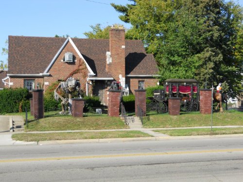 A brick house with a horse-drawn carriage and decorative figures in the front yard, surrounded by greenery.